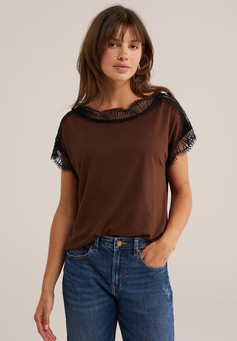 Jeune femme portant un haut marron avec bordure en dentelle noire et jean bleu, debout avec une main dans la poche sur fond uni.