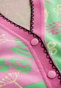 Gros plan sur un tissu tricoté rose pastel et vert avec des motifs floraux, une bordure noire en zigzag et deux boutons recouverts sur des bords côtelés.