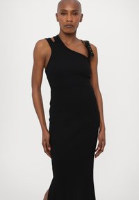 Black ribbed dress with sleeveless design, asymmetrical neckline, and gold-accented strap detail; fitted silhouette that reaches mid-calf.