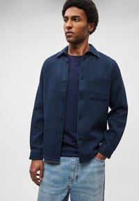 Mélykék texturált ingdzseki gombos elülső résszel, egyetlen mellkasi zsebbel és laza szabással, sötétkék ing és világos farmer fölött viselve.