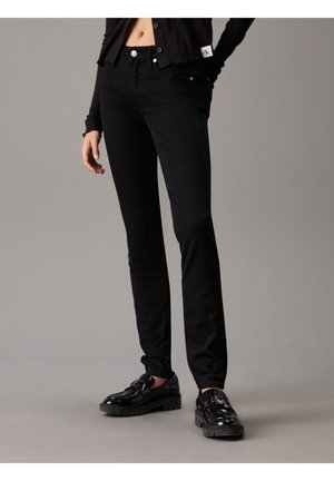 Person trägt schwarze Slim-Fit-Jeans, ein schwarzes geripptes, geknöpftes Oberteil und schwarze glänzende Slipper vor neutralem Hintergrund.