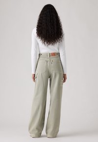 Γυναίκα με μακριά κατσαρά μαλλιά, που φορά λευκή μπλούζα με μακριά μανίκια και ανοιχτοπράσινο παντελόνι με φαρδιά πόδια, στέκεται με την πλάτη σε απλό φόντο.