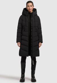 Abrigo acolchado negro con capucha, que presenta un cierre con cremallera, diseño acolchado y dos bolsillos laterales. Lleva puesto con botas negras hasta la rodilla.