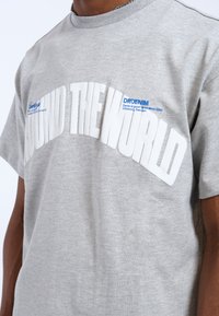 Camiseta de algodón gris con un audaz texto en relieve blanco que dice "AROUND THE WORLD" y una etiqueta de marca azul. Mangas cortas y escote redondo.