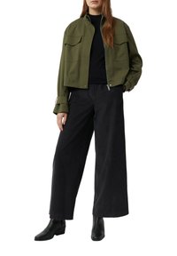 Frau in einer olivgrünen kurz geschnittenen Jacke, schwarzen weit geschnittenen Hosen, schwarzem Oberteil und schwarzen Knöchelstiefeln, die mit einer Hand in der Tasche steht.
