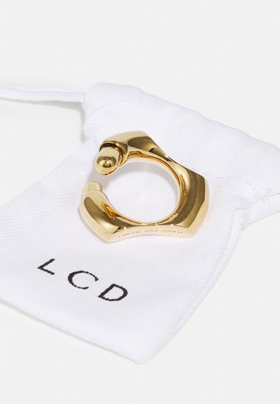 Gouden ring met een sculpturaal ontwerp, met een gladde textuur en ronde uiteinden, geplaatst op een witte stoffen zak met het opschrift "LCD."