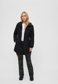 Svart parka-jakke med hette, to brystlommer og justerbar midje, kombinert med mørke camo-bukser og svart fottøy. Glatt tekstur.