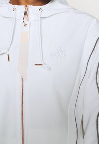 Vit zip-up huva jacka gjord av mjukt tyg, med roseguld hårdvara, ett präglat logotyp och tunna sidosträck.