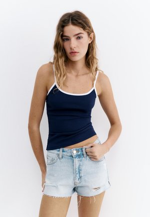 Ung kvinde med bølget hår iført en marineblå tanktop med hvide kanter og lyseblå slidte denimshorts, stående mod en hvid baggrund.