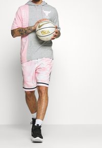 Grijze en roze hoodie met korte mouwen en logo; gecombineerd met roze mesh basketbalshorts met zwarte strepen; houdt een basketbal vast.
