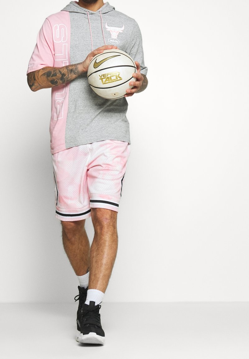 Grijze en roze hoodie met korte mouwen en logo; gecombineerd met roze mesh basketbalshorts met zwarte strepen; houdt een basketbal vast.