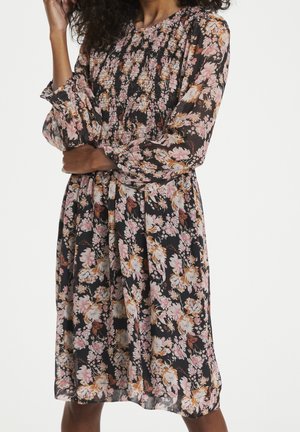 Kobieta ubrana w czarną, zwiewną sukienkę do kolan z różowo-beżowym kwiatowym wzorem i długimi bufiastymi rękawami, stojąca z założonymi ramionami.