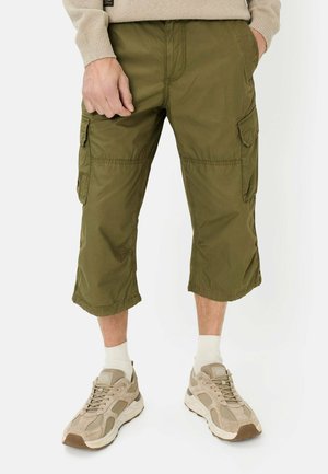 Mann trägt olivgrüne knielange Cargoshorts, beige Turnschuhe, weiße Socken und einen beigen Pullover, steht mit den Händen in den Taschen.