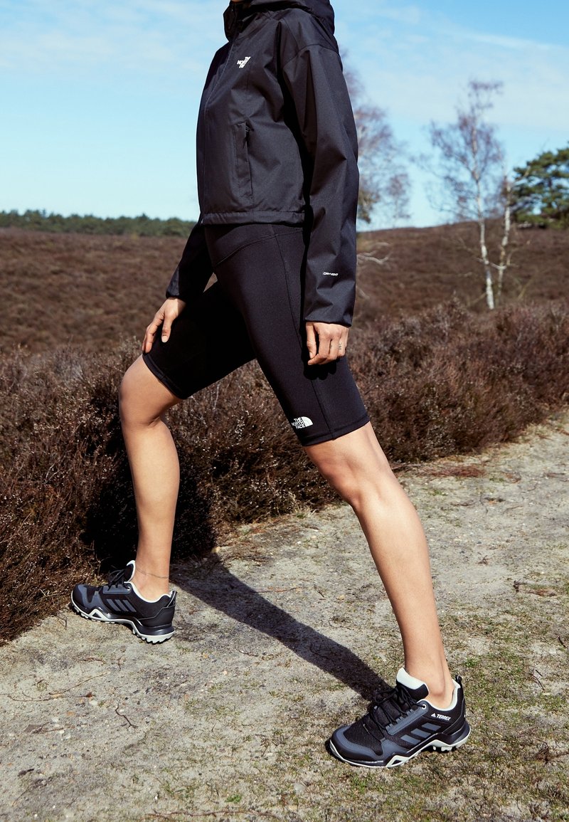 Sort vandtæt jakke med hætte, parret med sorte cykelshorts med et logo. Sorte sporty sko med en tekstureret sål og hvide detaljer.