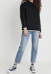 Sudadera negra con escote redondo, que presenta detalles y logo blancos en las mangas. Combinada con vaqueros de pierna recta en azul claro y zapatillas negras.