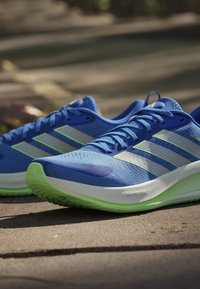 Paire de chaussures de course athlétiques bleues avec semelles vert clair et bandes blanches reposant sur un trottoir ensoleillé.