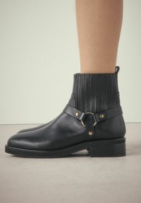 Botas de tobillo de cuero negro con panel acanalado texturizado, acento de hebilla de metal y suela plana. Cuenta con una lengüeta para facilitar su puesta.