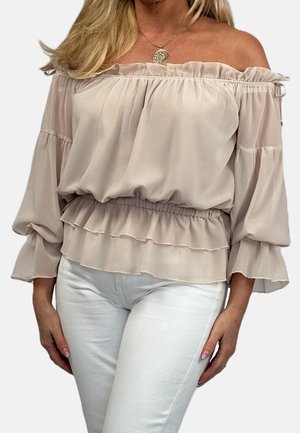 Femme portant un chemisier beige épaules dénudées avec des volants et des manches longues, associé à un pantalon blanc sur un fond uni.
