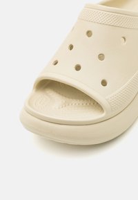 Beżowe klapki wykonane z teksturowanego plastiku, z otwartym palcem, dużymi otworami wentylacyjnymi i wyściełaną wkładką, charakteryzujące się gładkim wykończeniem.