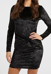 Robe en velours noir à manches longues, dotée d'un détail en front torsadé et d'une silhouette ajustée. Texture lisse avec un léger éclat.
