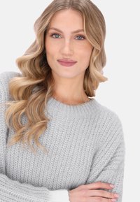 Jasnoszary sweter z dzianiny z okrągłym dekoltem i fakturowanym wzorem. Rękawy są białe. Modelka ma falowane włosy ułożone w delikatny sposób.