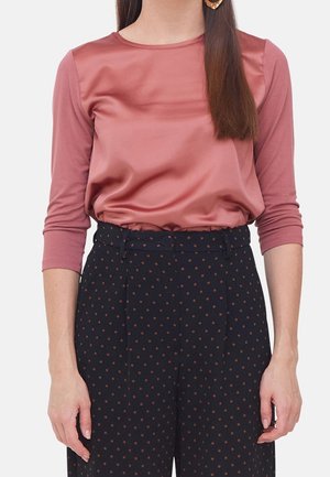 Femme portant un haut en satin rose poussiéreux à manches longues, rentré dans un pantalon noir taille haute à petits pois marron.