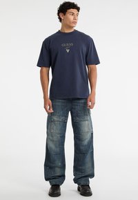 Man met krullend haar, gekleed in een marineblauwe Guess t-shirt, losse vervaagde cargo jeans en zwarte schoenen, staand tegen een witte achtergrond.