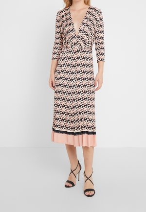 Femme portant une robe midi à motif géométrique rose et noir, manches trois-quarts, et talons hauts noirs à brides.