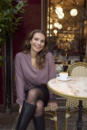 Nasmiješena žena u ljubičastoj bluzi i crnoj suknji sjedi za stolom na otvorenom kafiću s šalicom kave, okružena biljkama i prozorom.