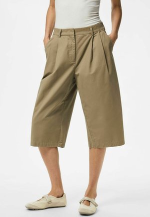 Persoon draagt losse, knie-lange beige culottes met zakken, gecombineerd met een wit topje en licht beige platte schoenen met bandjes.