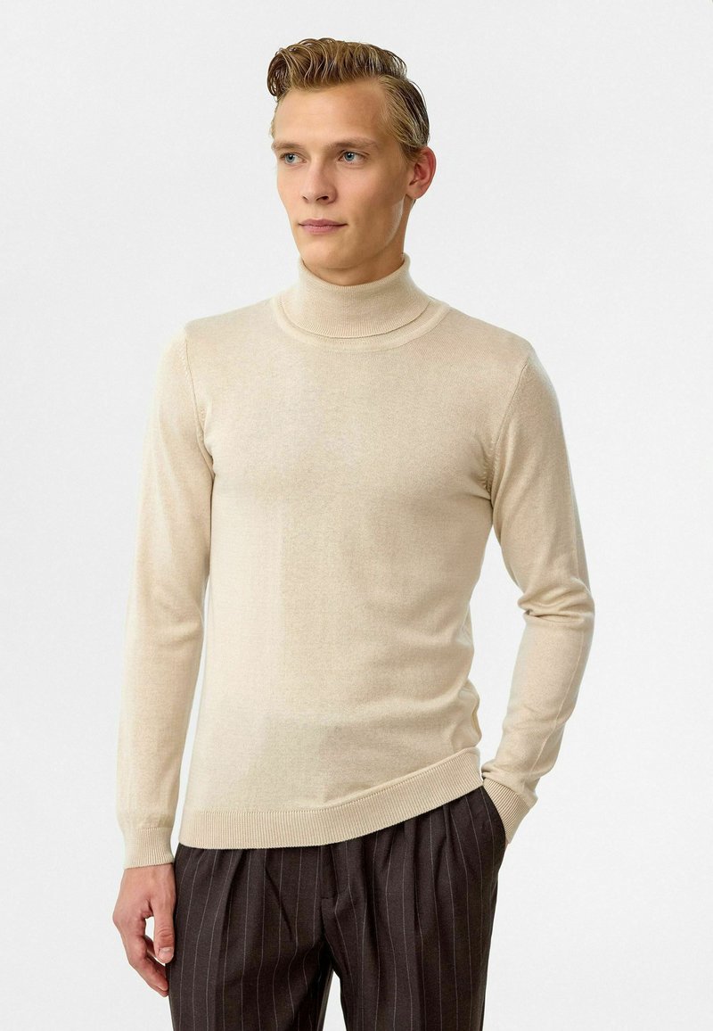 Beige coltrui met lange mouwen, fijn gebreide textuur, geribbelde kraag en zoom, gecombineerd met donker gestreepte pantalon.