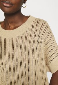 Musta ihoinen henkilö, joka käyttää kultaisia rengaskorviksia ja beigeä avoinneuleista lyhythihaista puseroa, jossa on ribbivarsi ja -kaulus.