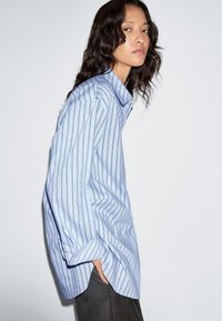 Light blue striped button-up shirt with a relaxed fit, featuring long sleeves and a pointed collar, paired with grey pants.