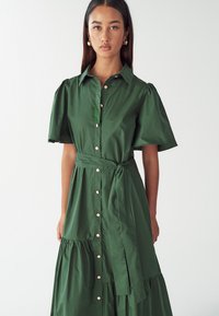 Zelené šaty na knoflíky vyrobené z hladké látky, s krátkými nafouknutými rukávy, stahovacím páskem a sukní se sklady a zlatými knoflíky vpředu.