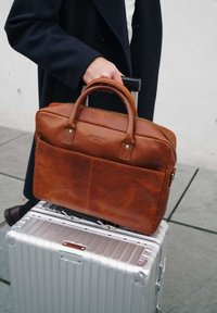 Brown leather laptop bag with sturdy handles, stitched accents, and a front pocket, placed atop a silver hard-shell suitcase.