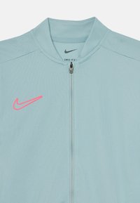 Veste zippée Nike Dri-FIT bleu clair avec un tissu texturé, un col haut, et un logo rose sur la poitrine gauche. Détail de fermeture éclair.