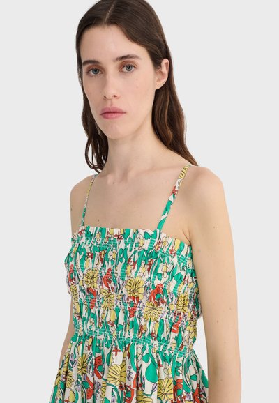 Vrouw met lang bruin haar, gekleed in een mouwloze, meerkleurige bloemenjurk met dunne bandjes en een geplooide taille, tegen een effen lichte achtergrond.