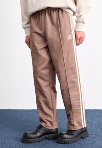 Brūnas sporta bikses ar baltām sānu joslām, izgatavotas no gludas auduma, ar elastīgu jostasvīru un Adidas logo kreisajā augšstilbā.