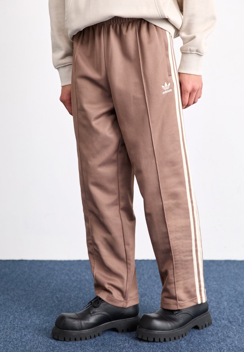 Brūnas sporta bikses ar baltām sānu joslām, izgatavotas no gludas auduma, ar elastīgu jostasvīru un Adidas logo kreisajā augšstilbā.
