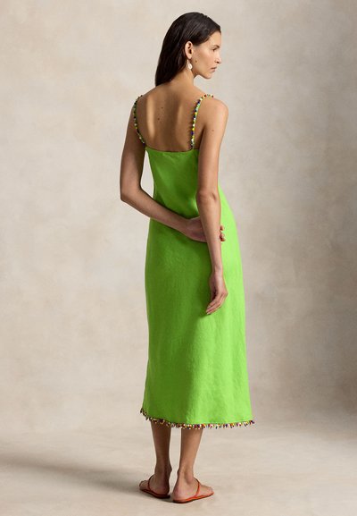 Nő háttal áll, lime zöld ujjatlan ruhát visel színes gyöngy díszítéssel és barna lapos szandált, egyszínű háttér előtt.