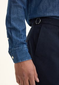 Dark blue denim shirt with buttoned cuff, paired with dark trousers featuring a silver buckle and detailed stitching along the waistband.