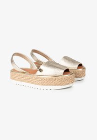 Zapatos slingback metálicos dorados con un diseño de punta abierta, superficie texturizada y suela de plataforma trenzada, que cuentan con una parte inferior de goma blanca para mayor agarre.