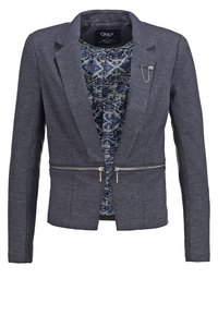 Blazer gris foncé sur mesure avec revers, doubles fermetures éclair à la taille, une doublure intérieure bleue à motifs, et une broche chaîne argentée sur le revers.