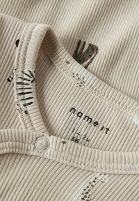 Tessuto beige a coste, con stampe animali scure. Chiusura a bottoni prominente al collo e bordi cuciti; etichetta del marchio visibile.