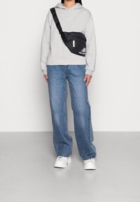 Bolso negro tipo bandolera con cremallera, acentos amarillos y logotipo; llevado sobre una sudadera gris y jeans acampanados azules, combinado con zapatillas blancas. 