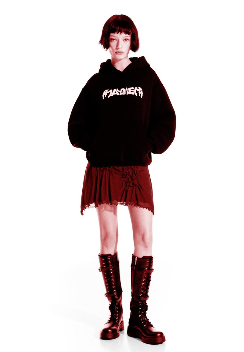 Črna kapuca z belim logotipom, rdeča plastena krila z čipkastim robom in črne vojaške škornje do kolen s vezalkami in zaponkami.