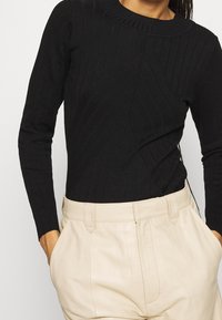 Person, der einen schwarzen gerippten Langarmpullover trägt, in hochgeschnittene, beigefarbene, gefaltete Hose gesteckt, mit Händen in den Taschen.