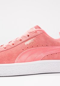 Różowe zamszowe sneakersy z tonującymi sznurowadłami, posiadające perforacje, teksturowaną białą podeszwę środkową oraz złote logo na boku.