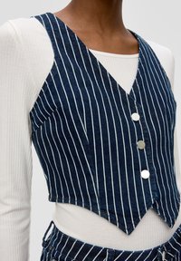 Denimweste mit V-Ausschnitt, die blaue und weiße vertikale Streifen aufweist, fünf silberne Knöpfe hat und im Crop-Design gestaltet ist. Getragen über einem weißen gerippten Oberteil.