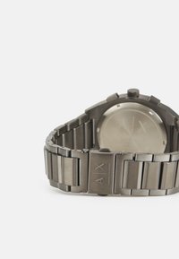Orologio in metallo con bracciale in acciaio inossidabile grigio, cassa rotonda e superficie liscia. Presenta un logo inciso sulla fibbia e un design minimalista.